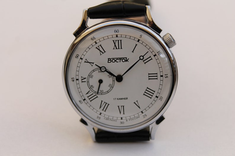 Puede incluir: Un reloj de pulsera plateado con una esfera blanca y marcadores de hora en n&uacute;meros romanos negros. El reloj tiene una correa de cuero negra y un peque&ntilde;o dial de segundero. La esfera del reloj tambi&eacute;n incluye el texto "BOCTOK" y "17 КАМНЕЙ".