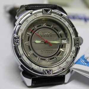 Puede incluir: Un reloj de pulsera plateado con una correa de cuero negro. La esfera del reloj es blanca con marcas negras y las palabras "Vostok" y "Rodika" impresas en ella. El reloj tiene una escala de tacómetro alrededor del borde exterior de la esfera.