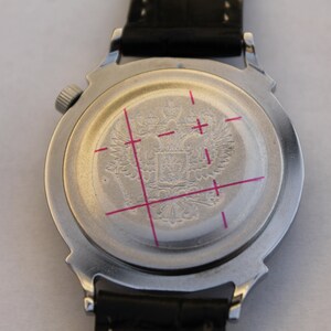 Puede incluir: La parte posterior de un reloj de pulsera plateado con una correa de cuero negro. La esfera del reloj tiene un dise&ntilde;o circular con un escudo de armas grabado que presenta un &aacute;guila bic&eacute;fala.