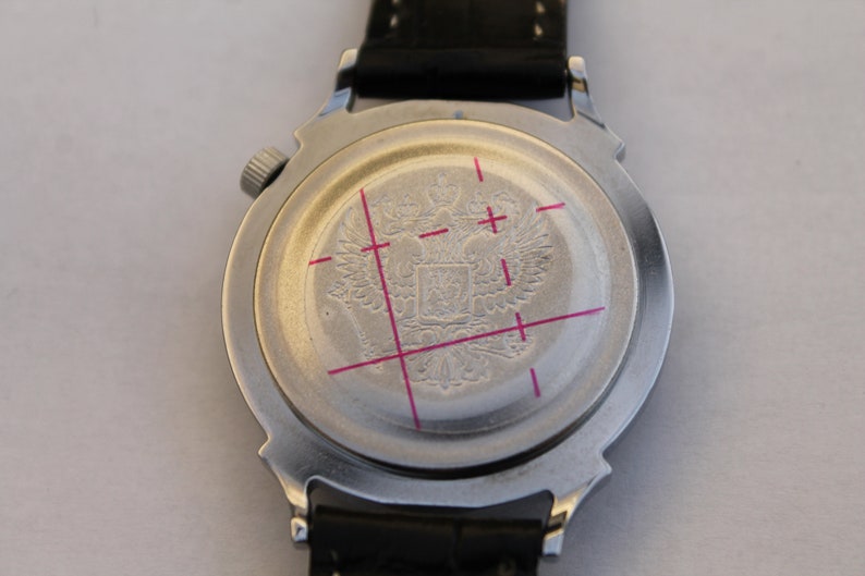 Puede incluir: La parte posterior de un reloj de pulsera plateado con una correa de cuero negro. La esfera del reloj tiene un dise&ntilde;o circular con un escudo de armas grabado que presenta un &aacute;guila bic&eacute;fala.