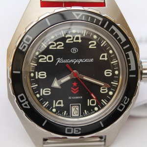 May include: A silver wristwatch with a black dial, white numerals, and hands. The watch has a black rotating bezel and a red second hand. The dial includes the text "Командирские" and "32 камня".