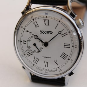 Puede incluir: Un reloj plateado con una esfera blanca y marcadores de hora en n&uacute;meros romanos negros. El reloj tiene una correa de cuero negro y un peque&ntilde;o dial de segundero. La esfera del reloj tambi&eacute;n incluye el texto "BOCTCK" y "17 KAMHEH".