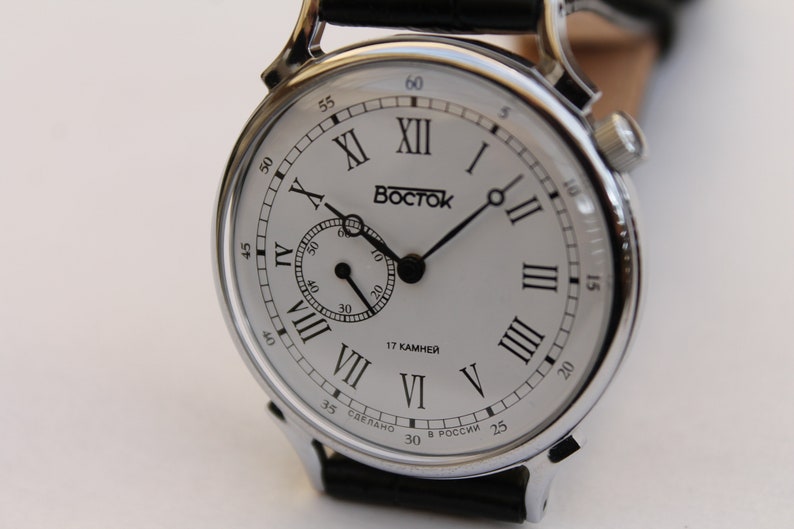 Puede incluir: Un reloj plateado con una esfera blanca y marcadores de hora en n&uacute;meros romanos negros. El reloj tiene una correa de cuero negro y un peque&ntilde;o dial de segundero. La esfera del reloj tambi&eacute;n incluye el texto "BOCTCK" y "17 KAMHEH".