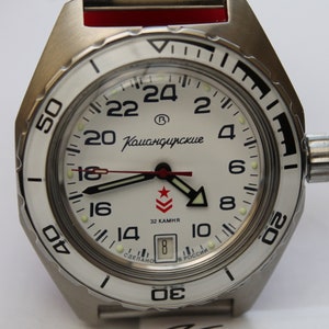 Può includere: Un orologio da polso con quadrante bianco con marcature nere e cinturino argentato. Il quadrante dell'orologio ha una lancetta dei secondi rossa, una lancetta delle ore nera e una lancetta dei minuti nera. Il quadrante dell'orologio ha anche una stella rossa e il testo "Командирские" in nero.