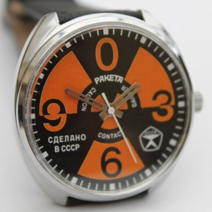 May include: A black and orange wristwatch with a circular face. The face has a large orange radiation symbol with black numbers 0, 3, 6, and 9. The watch face also has the text "RAKETA", "CAUTION", "СДЕЛАНО В СССР", and "CONTACT".