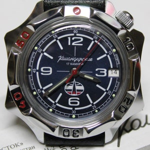 May include: A silver wristwatch with a black face and red accents. The watch face has white markings and the text "Командирские 17 КАМНЕЙ".