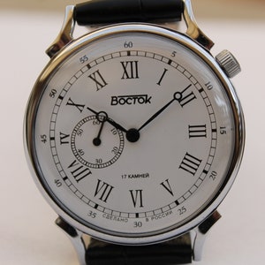 Puede incluir: Un reloj de pulsera plateado con una esfera blanca y marcadores de hora en n&uacute;meros romanos negros. El reloj tiene una correa de cuero negro y un peque&ntilde;o dial de segundero. La esfera del reloj tambi&eacute;n incluye el texto "Восток" y "17 КАМНЕЙ".