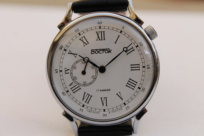 Puede incluir: Un reloj de pulsera plateado con una esfera blanca y marcadores de hora en n&uacute;meros romanos negros. El reloj tiene una correa de cuero negro y un peque&ntilde;o dial de segundero. La esfera del reloj tambi&eacute;n incluye el texto "Восток" y "17 КАМНЕЙ".
