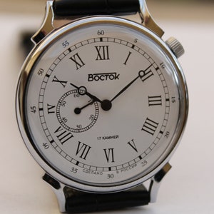 Puede incluir: Un reloj plateado con una esfera blanca y marcadores de hora en n&uacute;meros romanos negros. El reloj tiene una correa de cuero negro y dice "BOSTOK" y "17 КАМНЕЙ" en la esfera.