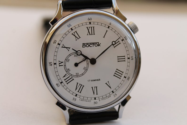 Puede incluir: Un reloj plateado con una esfera blanca y marcadores de hora en n&uacute;meros romanos negros. El reloj tiene una correa de cuero negro y dice "BOSTOK" y "17 КАМНЕЙ" en la esfera.