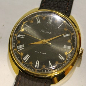 May include: A gold-tone wristwatch with a black face and Roman numeral hour markers. The watch has a brown leather strap and says "Raketa" and "Made in USSR" on the face.