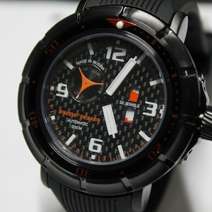 Könnte beinhalten: Eine schwarze Armbanduhr mit einem orangefarbenen und weißen Zifferblatt. Das Zifferblatt der Uhr hat ein Karbonfaser-Muster und den Text "AMFIBIA AUTOMATIC 200 M" und "MADE IN RUSSIA".