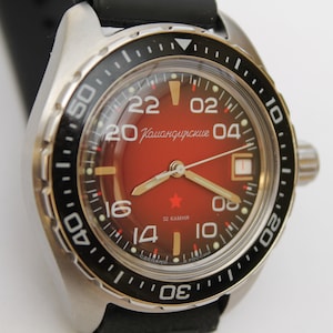 May include: A red and silver men's wristwatch with a black bezel and a red face. The face has white numbers and a red star in the centre. The watch has the text "Командирские" on the face.