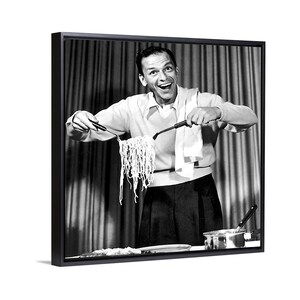 May include: Black and white framed print of a man holding up a large portion of spaghetti with tongs. The man is wearing a light-colored sweater and dark pants. A white towel is draped over his arm.