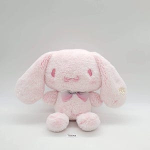 Könnte beinhalten: Ein rosa Plüschtier-Hase mit langen, schlaffen Ohren, einer kleinen rosa Schleife und einem aufgenähten Gesicht. Der Hase hat eine flauschige Textur und sitzt aufrecht. Das Spielzeug befindet sich auf weißem Hintergrund.