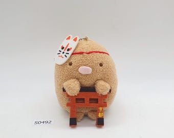 May include: A plush toy in the shape of a light brown, rounded character, wearing a white fox mask and holding a small, orange torii gate. The gate has black accents and Japanese text. The toy has a red headband.