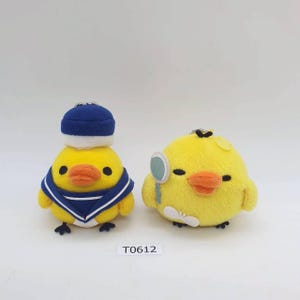 May include: Two plush yellow chick keychains. One chick wears a blue sailor hat and a blue and white sailor collar. The other chick holds a magnifying glass. Both have orange beaks and black eyes. The text "T0612" is below the chicks.