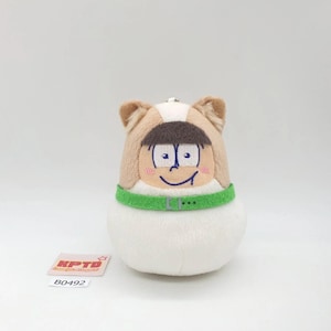 May include: A plush toy shaped like a rounded figure with tan and white fur, featuring embroidered facial details, a brown bowl-cut hairstyle, and a bright green collar. A small rectangular tag with the text KPTD and B0492 is placed nearby.