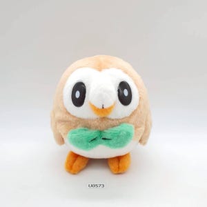 Op de afbeelding: Een pluche uil speelgoed, in lichtbruin en wit, met grote zwarte ogen en een oranje snavel. Het heeft een groene vlinderdas en oranje voeten. De productcode "U0573" is onderaan afgedrukt. Een kinderspeelgoed.