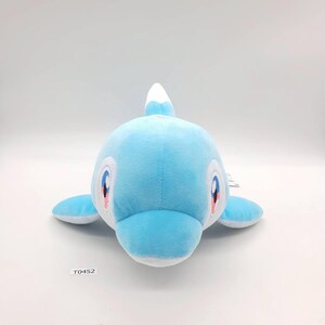 May include: A light blue plush dolphin toy with white detailing. The dolphin features large, friendly eyes with pink and blue accents. It has a rounded body, small fins, and a white dorsal fin. The tag reads "T0452".