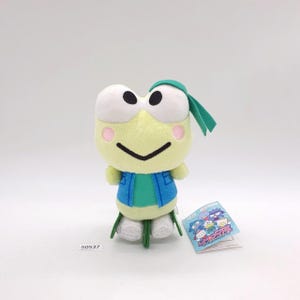 May include: A plush frog toy with a light green body, white eyes, and pink cheeks. It wears a blue vest and a green headband. A tag with Japanese text is attached. The toy is approximately 5 inches tall.