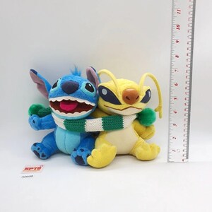 Puede incluir: Dos peluches, uno azul y otro amarillo, envueltos en una bufanda de punto verde y blanca. Una regla vertical muestra una altura de aproximadamente 15 cm. El juguete azul tiene orejas grandes y boca abierta, el amarillo tiene antenas largas.
