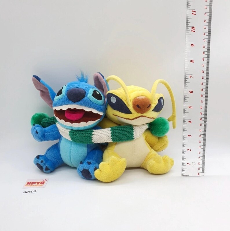 Puede incluir: Dos peluches, uno azul y otro amarillo, envueltos en una bufanda de punto verde y blanca. Una regla vertical muestra una altura de aproximadamente 15 cm. El juguete azul tiene orejas grandes y boca abierta, el amarillo tiene antenas largas.