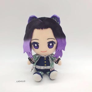Könnte beinhalten: Plüschpuppe eines Anime-Charakters mit schwarzen und lila Haaren, großen Augen und einem lächelnden Gesichtsausdruck. Die Puppe trägt ein marineblaues Outfit mit weißem Gürtel und einer gemusterten Jacke. Die Puppe sitzt.