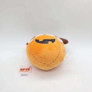 K&ouml;nnte beinhalten: Ein rundes, orangefarbenes Pl&uuml;schtier mit einem schwarz gestickten Mund und braunen Ohren. Das Pl&uuml;schtier hat dekorative braune Ziern&auml;hte auf der Oberfl&auml;che. Daneben liegt ein kleines wei&szlig;es quadratisches Etikett mit dem Text KPTD und A0504.