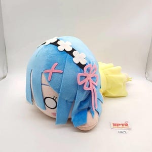 May include: A plush toy featuring a character with light blue hair, a black headband with white flower accents, and a pink bow. The character has large eyes and a light skin tone. A small white rectangular tag with the text KPTD L0679 is visible.