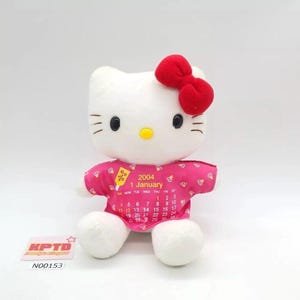 Puede incluir: Un peluche blanco con un lazo rojo, ojos negros y nariz amarilla. Viste una camisa rosa con un calendario de enero de 2004. El juguete tiene pies y brazos blancos. Una etiqueta con el texto "KPTD" y "N00153" es visible.
