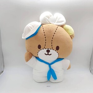 May include: A plush brown bear wearing a white sailor outfit with a blue tie and hat. The bear has a white bow on its head and a stitched face. The bear is a stuffed animal.