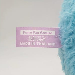 May include: A close-up of a light purple tag with the text "Fun Fan Amuse" and "SEGA" in white. The tag is attached to a fluffy, light blue plush toy. The tag also reads "MADE IN THAILAND".