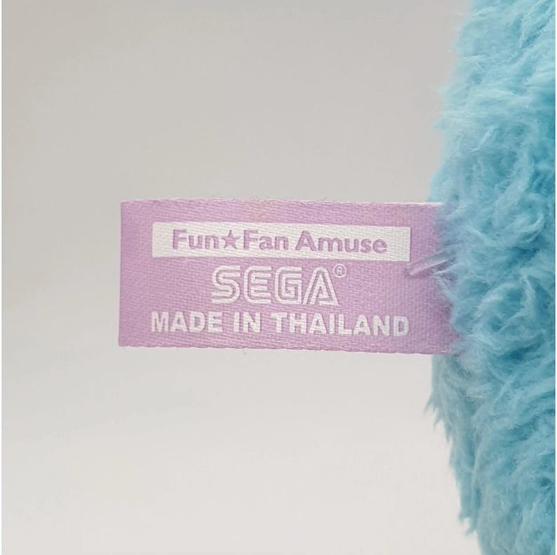 May include: A close-up of a light purple tag with the text "Fun Fan Amuse" and "SEGA" in white. The tag is attached to a fluffy, light blue plush toy. The tag also reads "MADE IN THAILAND".