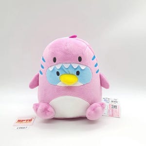 May include: A pink plush toy shaped like a penguin with a shark-like design. The toy has a blue face with white teeth, a yellow beak, and blue stripes. It has small arms and feet, and a white belly.