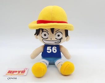 May include: A plush doll of a character wearing a yellow straw hat with a red band, a blue tank top with the number 56, and yellow shorts. The doll has black hair and a stitched face with a scar. A tag with the text "KPTD" and "Q0496" is visible.