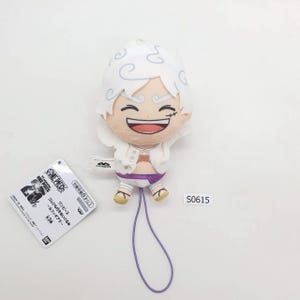May include: A One Piece anime character plush toy. The figure has white hair, a smiling face, and is wearing a white jacket and purple shorts. A tag with Japanese text is attached. The code S0615 is visible.
