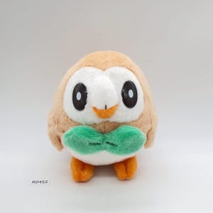 May include: A plush owl toy with a tan body, white face, and large black eyes. It has a green bow tie and orange feet. The toy is on a white background. The text "R0455" is visible.
