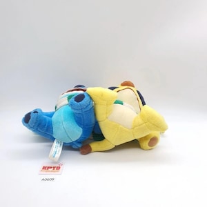 Puede incluir: Dos juguetes de peluche acostados de lado, uno azul y otro amarillo, ambos con detalles azul oscuro en las orejas y los pies, una mancha de color crema en el pecho y una bufanda a rayas verdes y blancas alrededor del cuello, con una etiqueta.