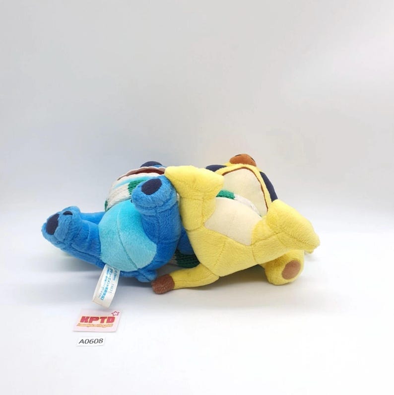 Puede incluir: Dos juguetes de peluche acostados de lado, uno azul y otro amarillo, ambos con detalles azul oscuro en las orejas y los pies, una mancha de color crema en el pecho y una bufanda a rayas verdes y blancas alrededor del cuello, con una etiqueta.