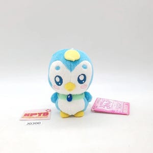 Puede incluir: Un pequeño peluche con forma de pingüino azul y blanco con pico y pies amarillos, un collar verde claro con una gema ovalada azul oscuro y marcas de rubor rosa en las mejillas. Se acompaña de una etiqueta rosa y una tarjeta blanca con KPTD J0300.