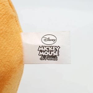 K&ouml;nnte beinhalten: Eine weiche, goldgelbe Pl&uuml;schstoffoberfl&auml;che mit einem wei&szlig;en rechteckigen Etikett. Das Etikett zeigt oben das Disney-Logo, gefolgt von dem schwarzen Text MICKEY MOUSE & Friends, der in einer fetten, stilisierten Schrift auf wei&szlig;em Grund gedruckt ist.