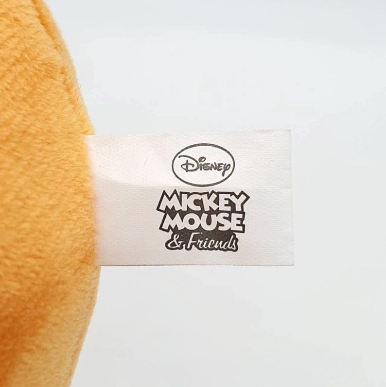 K&ouml;nnte beinhalten: Eine weiche, goldgelbe Pl&uuml;schstoffoberfl&auml;che mit einem wei&szlig;en rechteckigen Etikett. Das Etikett zeigt oben das Disney-Logo, gefolgt von dem schwarzen Text MICKEY MOUSE & Friends, der in einer fetten, stilisierten Schrift auf wei&szlig;em Grund gedruckt ist.