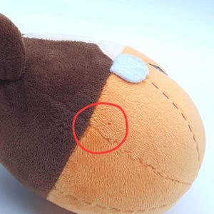 K&ouml;nnte beinhalten: Ein Pl&uuml;schtier mit einer braun-orangen Stoffoberfl&auml;che und sichtbaren N&auml;hten. Ein roter Kreis markiert eine kleine Stelle mit Abnutzung oder Besch&auml;digung auf dem orangefarbenen Bereich des Pl&uuml;schmaterials. Das Spielzeug hat eine weiche Textur.