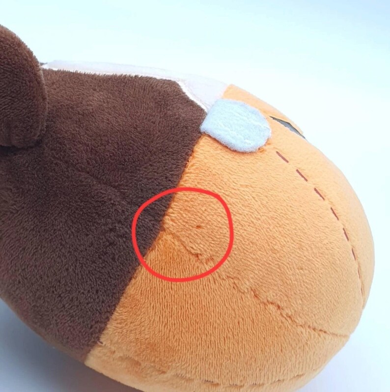 K&ouml;nnte beinhalten: Ein Pl&uuml;schtier mit einer braun-orangen Stoffoberfl&auml;che und sichtbaren N&auml;hten. Ein roter Kreis markiert eine kleine Stelle mit Abnutzung oder Besch&auml;digung auf dem orangefarbenen Bereich des Pl&uuml;schmaterials. Das Spielzeug hat eine weiche Textur.