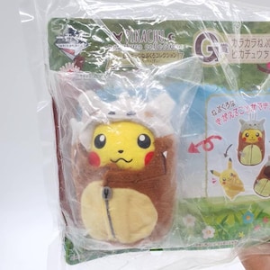 May include: A Pikachu plush toy in a brown outfit, inside a clear plastic package. The plush has a yellow face with black eyes and red cheeks. The package has Japanese text and the Pikachu logo.