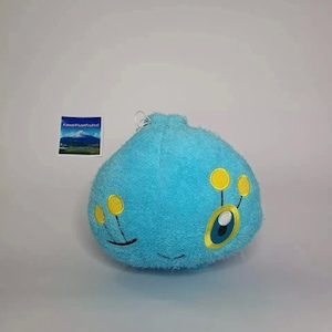 May include: A bright blue plush toy shaped like a rounded blob with a fuzzy texture. It features two yellow antennae with circular tips and a single large eye with a blue iris and white pupil. A small square tag displays a mountain landscape image.