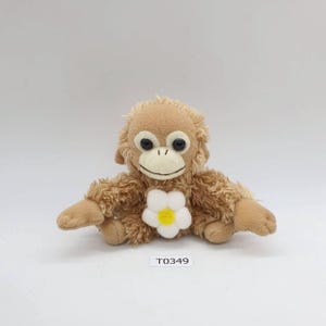 Może przedstawiać: Pluszowa brązowa zabawka małpka z dużymi czarnymi oczami, wyszywanym uśmiechem i białym kwiatem z żółtym środkiem na piersi. Zabawka ma wyciągnięte ramiona, a na metce napis "T0349".