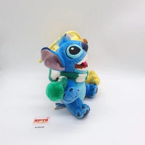 Puede incluir: Un juguete de peluche azul con orejas grandes, que lleva una bufanda de punto verde y blanca con un pomp&oacute;n verde. El personaje tiene ojos negros, una lengua rosa y una peque&ntilde;a etiqueta en el pie. Se ve una etiqueta cuadrada blanca con KPTD A0608.