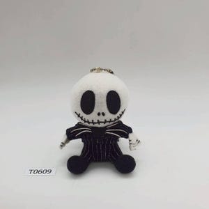 May include: A soft toy of Jack Skellington from The Nightmare Before Christmas. The figure has a white head with large black eyes and a stitched smile. It wears a black pinstripe suit and has a silver keychain attached. The tag reads T0609.